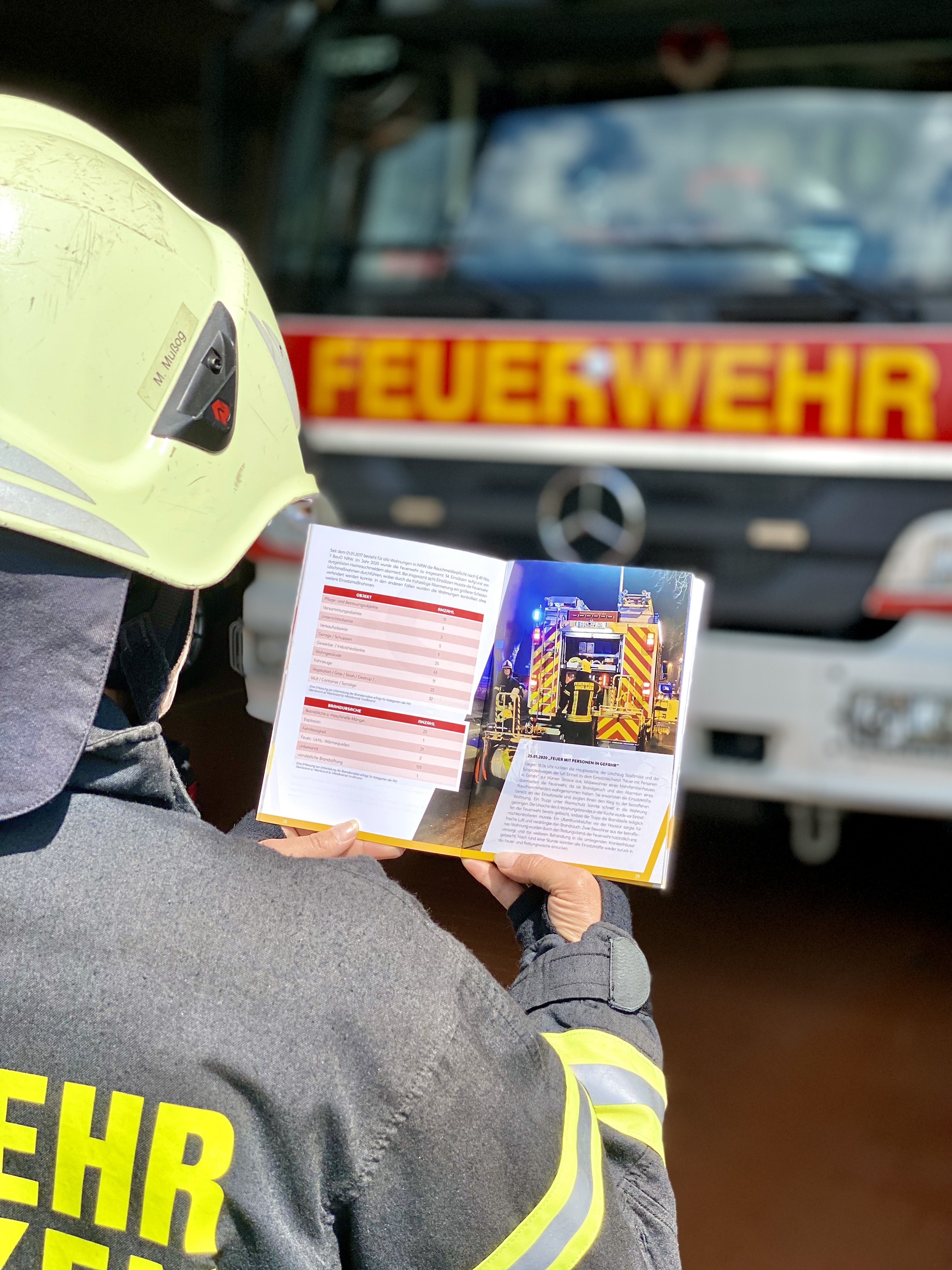 Feuerwehrfrau mit Jahresbericht in der Hand und Einsatzfahrzeug im Hintergrund